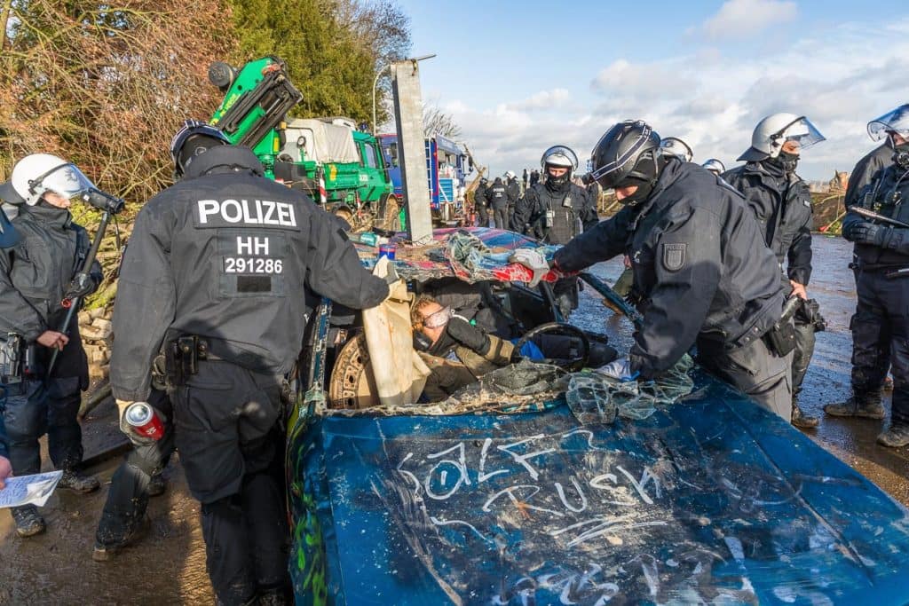 Polizisten versuchen, einen Klimaaktivisten aus einem Autowrack zu entfernen, in dem er sich zusammen mit einem Kanaldeckel festgeklebt hat. Das Autowrack ist mit einem stabilen Pfeiler als Barrikade auf einer Zufahrtsstraße nach Lützerath fixiert / © Foto: Georg Berg