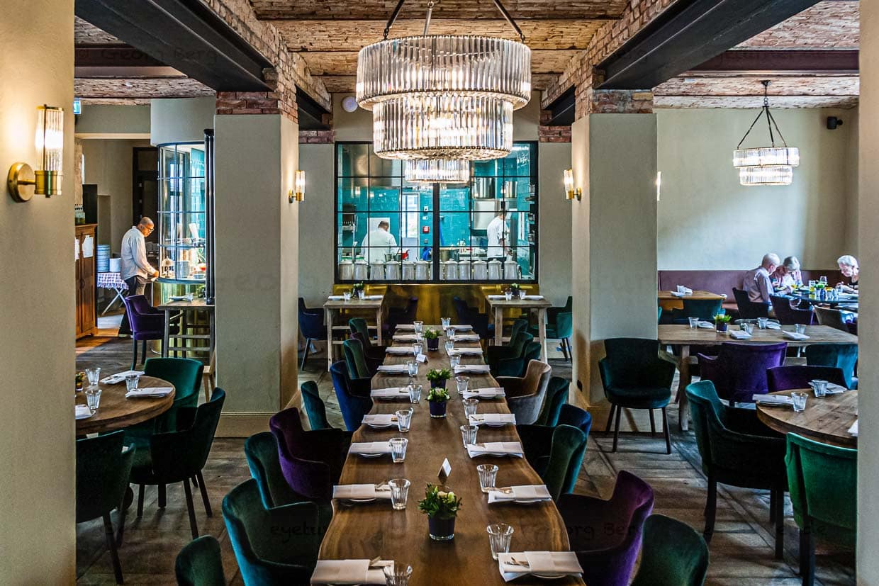 In der Mitte des Restaurants steht ein „Table Commune“ mit 20 Sitzplätzen. Er bleibt in der Regel frei für Gäste ohne Reservierung. Eine schöne Gelegenheit mit fremden Menschen ins Gespräch zu kommen. Schon der Raum mit seiner Architektur und dem Blick in die Küche bietet genügend Gesprächsansätze! / © Foto: Georg Berg