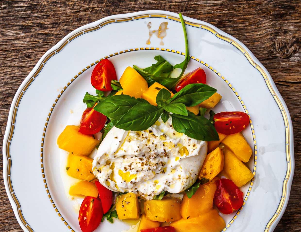 Pudel mit Goldrand auf dem Restaurantgeschirr. Leuchtend gelb in der Mitte ein wunderbares Sommergericht: Burrata mit Mango-Minz-Salat. Effektvoll gewürzt mit Olivenöl, crunchigem Meersalz und weißem Pfeffer / © Foto: Georg Berg