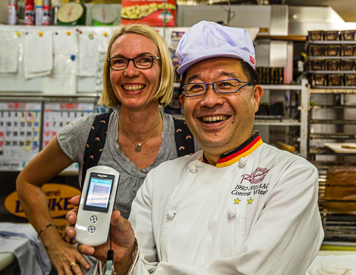 Daiichi Sugiyama trägt eine Bäckerjacke mit schwarz-rot-goldenen Streifen und benutzt virtuos einen mobilen Sprachübersetzer / © Foto: Georg Berg