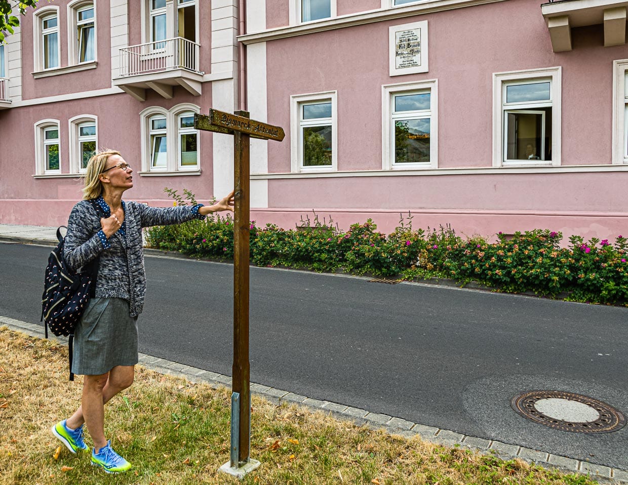 Panneau de signalisation de l'assassinat de Bismarck. Un panneau indicateur en bois et une plaque apposée sur la maison rappellent le lieu de l'assassinat de Bismarck en 1874. Le médecin traitant de Bismarck habitait dans cette maison rose située directement au bord du Rosengarten. Au petit matin, Bismarck partit de là en direction de la saline supérieure pour une inhalation / © Foto : Georg Berg