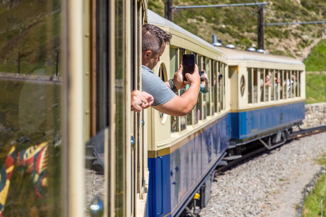 Voyageurs en train dans l'Alpine Classic Pullman Express au col de l'Oberalp à 2.000 mètres d'altitude photographient depuis le wagon lors du voyage nostalgique de Davos à Andermatt / © Foto : Georg Berg