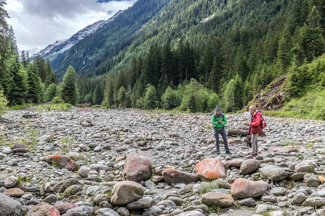Zwei Wanderer erkunden das steinige Flussbett am Wasserfallrundweg Untersulzbach, Rosental, Austria, umgeben von bewaldeten Bergen und schneebedeckten Gipfeln