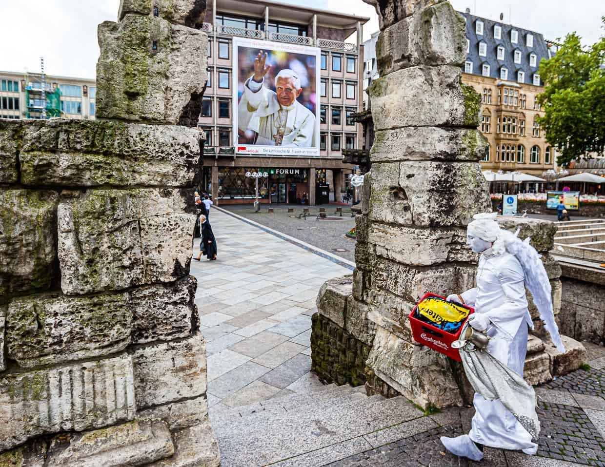Ángel con caja de cerveza ante el Papa. Una estatua viviente atraviesa la Puerta Romana frente a la catedral de Colonia con una caja de cerveza, que utilizará como pedestal. Al fondo, el Papa Benedicto VI, más grande que la vida, saluda desde un cartel / © Foto: Georg Berg