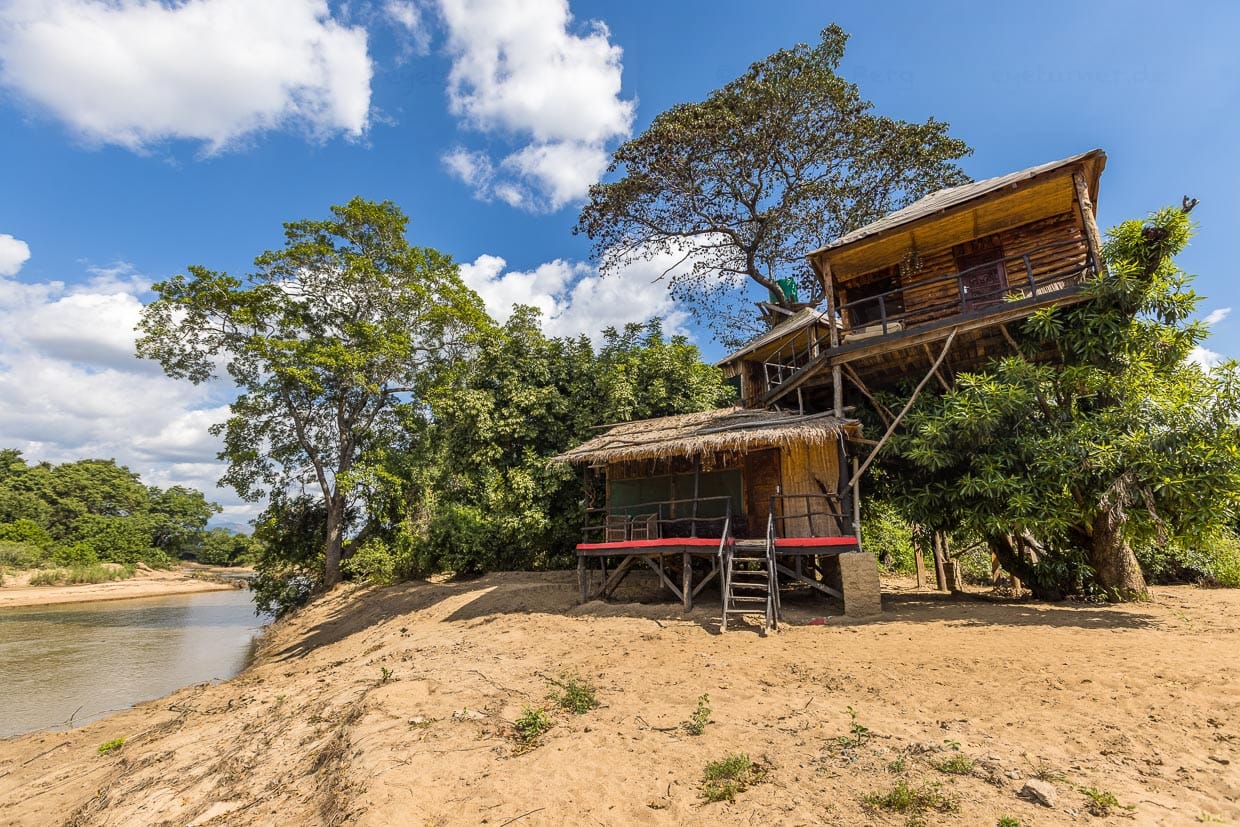 Baumhaus-Ensemble direkt am Likwenu Fluss. Hinter den Häusern ragt ein alter Mahagonibaum hervor. Im Fluß leben Krokodile und Fluspferde / © Foto: Georg Berg