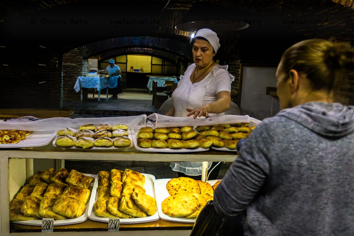 Die besten Backwaren von Tbilisi findet man unter dem Sophiko Chiaureli Garten. Eine Frau in weißer Kleidung und Kopftuch steht hinter einer Theke und bietet Backwaren an. Zahlreiche Backwaren liegen auf Tabletts in der Auslage vor einer Person. Im Hintergrund ist eine weitere Person zu sehen. Tbilisi, Georgien. / © Foto: Georg Berg