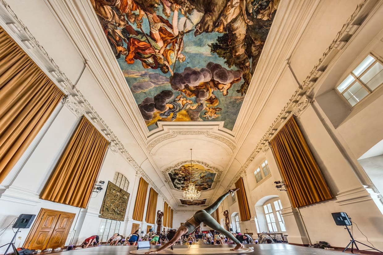 Eine Yogalehrerin leitet eine Gruppe beim Yoga in einem historischen Saal mit bemalter Decke am Domplatz, Salzburg, Austria