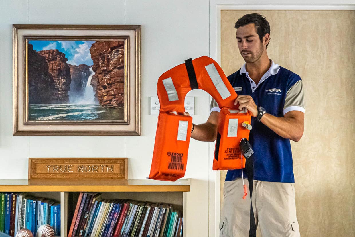 Jaydon, der erste Offizier an Bord, erklärt beim Sicherheitsbriefing, warum man beim Sprung ins Wasser die angelegte Schwimmweste mit den Händen umklammern soll: „Ihr könntet euch sonst auf die Zunge beißen. Aber merkt euch ruhig mal, dass ihr auch mit abgebissener Zunge immer noch in die Notfallpfeife blasen könnt“ / © Foto: Georg Berg