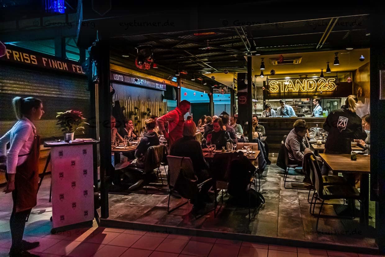 Wie eine warme, leuchtende Insel der Glückseligkeit. Wenn die Marktstände schon längst geschlossen sind, wird es erst richtig munter im Stand 25. Wer für Abends einen Tisch reserviert, wird an der Eingangspforte der ehrwürdigen Markthalle an der Stand Utca abgeholt / © Foto: Georg Berg