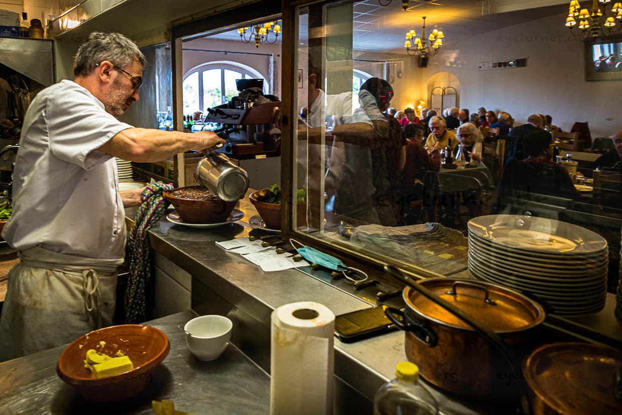 Chef Eric Rousselot bereitet das berühmte Cassoulet Imperial in der Hostellerie Etienne zu, die seit 1956 als Familienbetrieb geführt wird. Dicht an dicht sitzen die Gäste im Restaurant Etienne auch an einem Wochentag im November. Eric Rousselot gießt noch etwas Boullion an, bevor die nächste Cassole an den Tisch geht / © Foto: Georg Berg