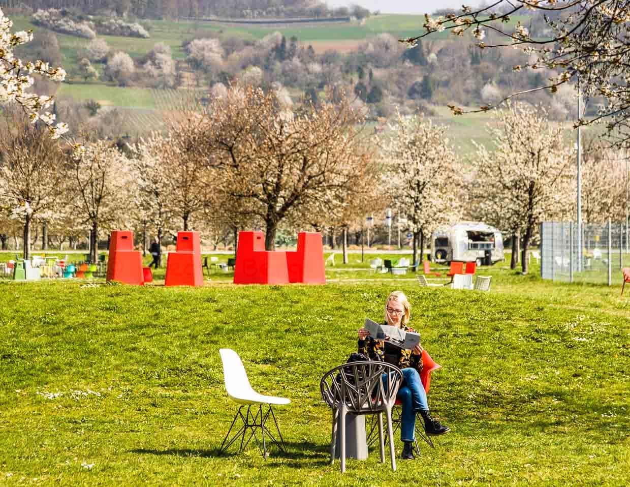 Willkommen in der Stadt der Stühle. Auf der Wiese vor dem Vitra Haus stehen unzählige Designer Stühle in der Landschaft. Als Besucher rückt man sich den Panton, Eames oder einen Vegetal von Bouroullec zurecht. Im Hintergrund eine Gruppe von roten Stool Tools von Konstantin Grcic. Noch weiter dahinter die Weinberge von Weil / © Foto: Georg Berg