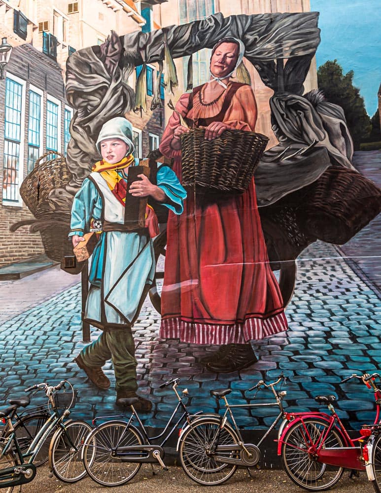 Wandmalerei in der Altstadt von Deventer. Motiv anlässlich des Hansejahres, das die neun holändischen Hansestädte 2023 feiern. Die Wandmalerei hat einen Bezug zur Vergangenheit und Gegenwart. Hier in Deventer: Marktfrau mit Sohn verkauft Deventer Honigkuchen und Stockfisch, wichtige Waren im Hansehandel von Deventer. Der Junge trägt einen Fan-Schal des Fußball-Clubs Deventer Eagle / © Foto: Georg Berg