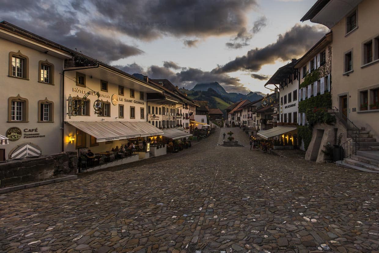 Hauptstraße der mittelalterlichen Stadt Gruyères mit vielen Restaurants, in denen die Spezialität Käsefondue Moité-Moité angeboten werden. Schloss St. Germain und Schloss Gryerz liegen in hinteren Teil des kleinen Ortes, der 2021 von der UNWTO mit dem Titel „Best Tourism Villages“ ausgezeichnet / © Foto: Georg Berg