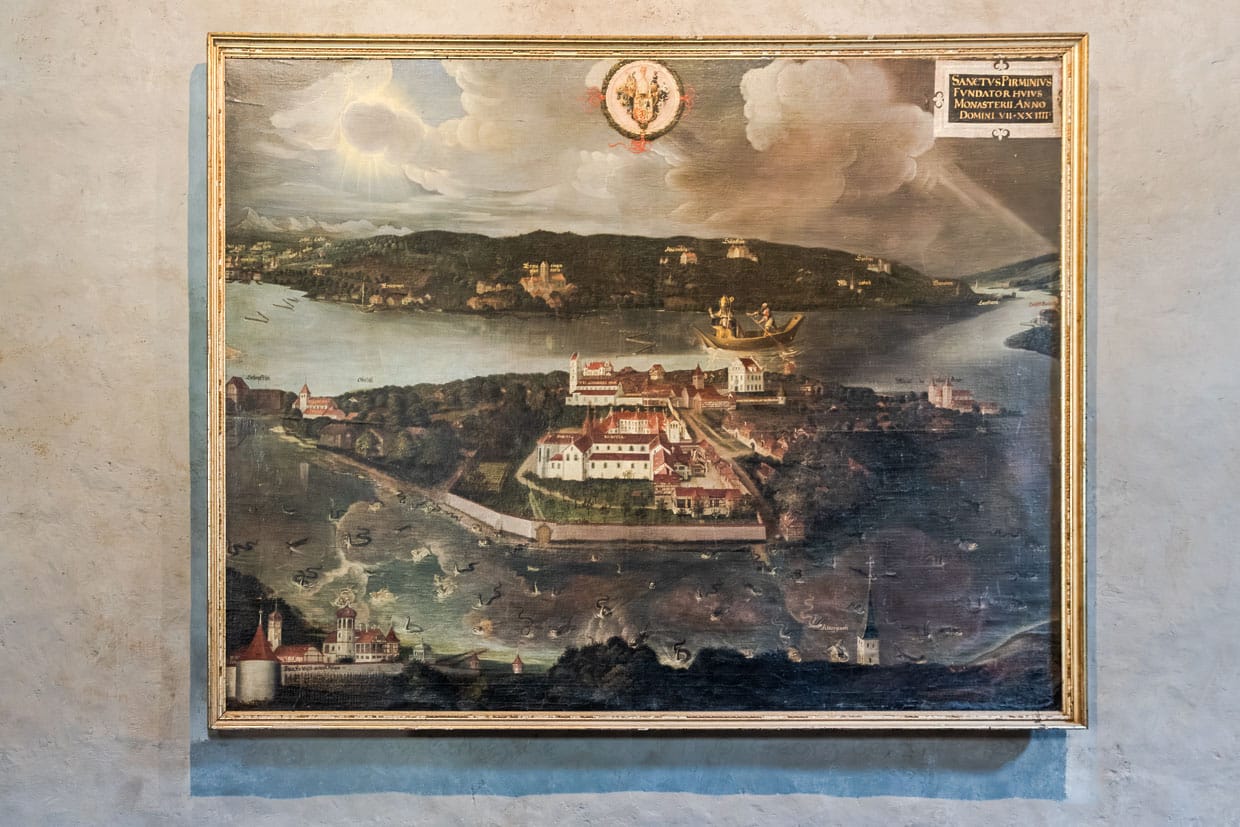 Das Tafelgemälde  von 1624 im Münster St. Maria und Markus veranschaulicht die Gründungslegende der Insel Reichenau und zeigt die Ankunft des Heiligen Pirmin. Die Bildmitte nimmt die ummauerte Klosteranlage mit der im 19. Jh. abgerissenen Abtspfalz und der Pfarrkirche St. Johann ein. Direkt am Hafen steht das „Hohe Haus“, am linken Bildrand sind die Ruine Schopflen und St. Georg in Oberzell, am rechten Bildrand St. Peter und Paul in Niederzell zu erkennen / © Foto: Georg Berg