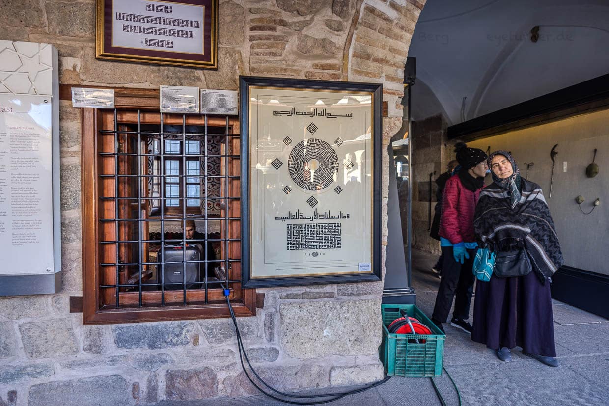 Hinter dem vergitterten Fenster eines traditionellen Derwisch-Raumes sitzt ein junger Mann. Über eine Lautsprecheranlage ertönen seine Koranverse über den Innenhof des Mevlana Museums / © Foto: Georg Berg