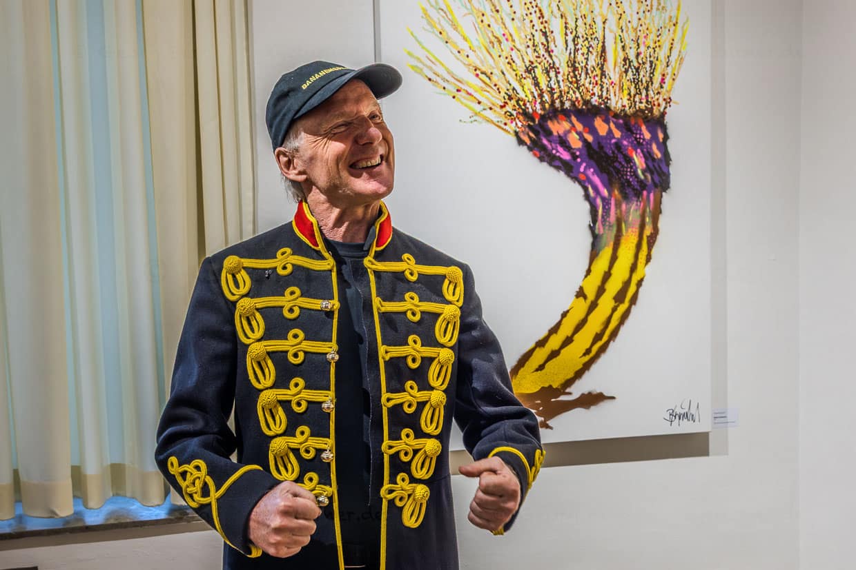 Ein lächelnder Mann in einer historischen Uniformjacke und Cap steht vor einem Kunstwerk auf der Schanze in Grevenbroich, Deutschland