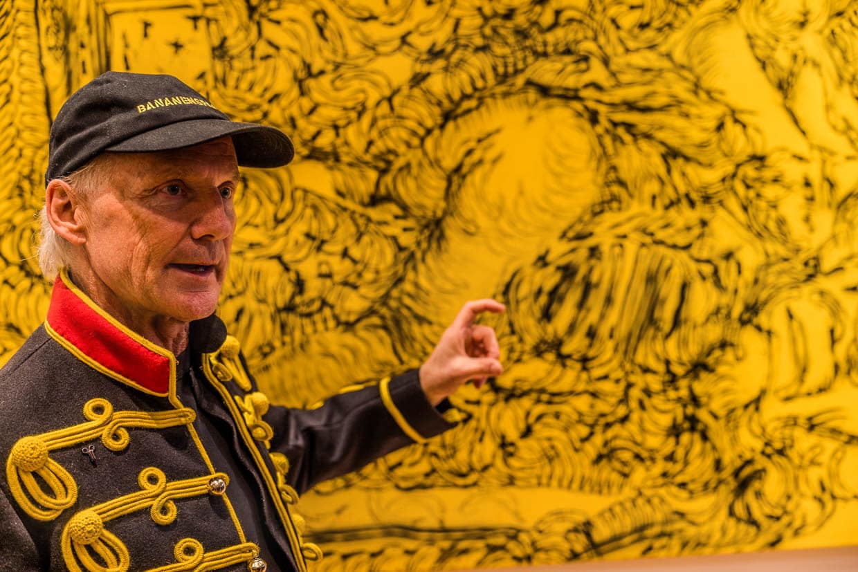 Mann mit Bananensprayer-Kappe präsentiert ein Kunstwerk vor gelbem Hintergrund am Stadtpark, Grevenbroich, Deutschland