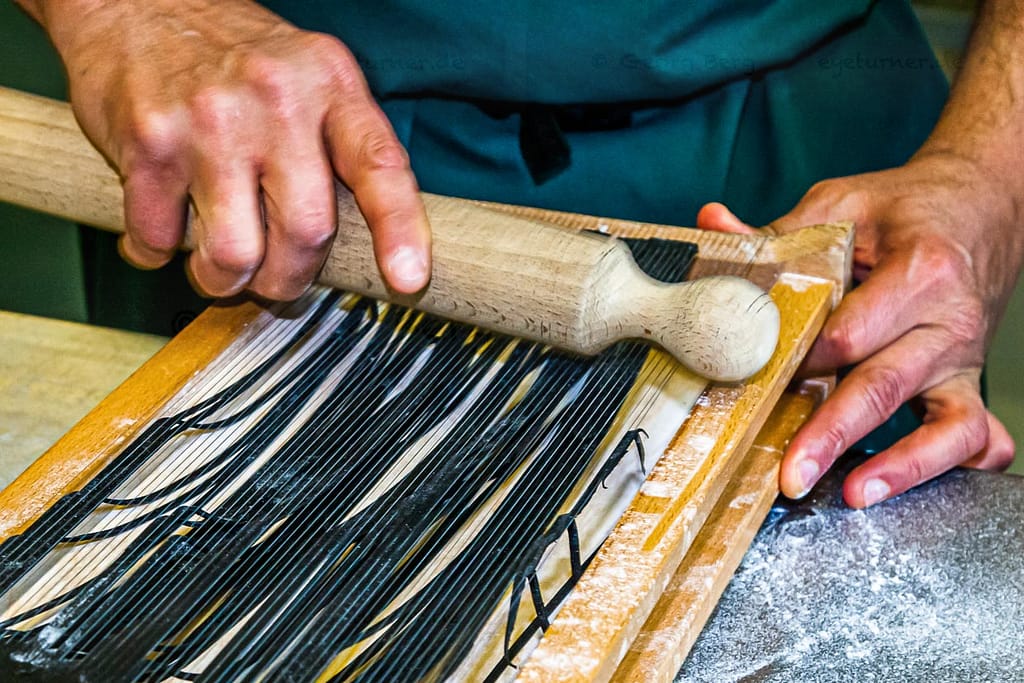 Die einzelnen Schritte der Pastaproduktion mit dem Carraturo. Zuerst wird der Teig mehrmals durch eine Nudelmaschine gedreht, bis er ganz dünn ist. Danach kann der dünne Teiglappen mit einem Nudelholz durch die Drähte gedrückt werden, die ihn in dünne Streifen schneiden. Anschließend kurz bemehlen und erst kurz vor dem Verzehr in Salzwasser al dente kochen. An der Seite des Chefkochs steht Salvatore Piacente, ebenfalls Absolvent der Kochschule Santa Maria, an der schon José Graziosi sein Handwerk lernte / © Foto: Georg Berg