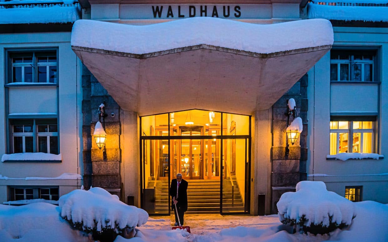 Meterhoher Neuschnee vor dem Waldhaus in Sils wird schon früh morgens weggeräumt. Das Licht in der Lobby leuchtet warm und einladend. Schön zu wissen, dass man sich gleich in einen der Samtsessel im Salon-Bleu fallen lassen kann / © Foto: Georg Berg
