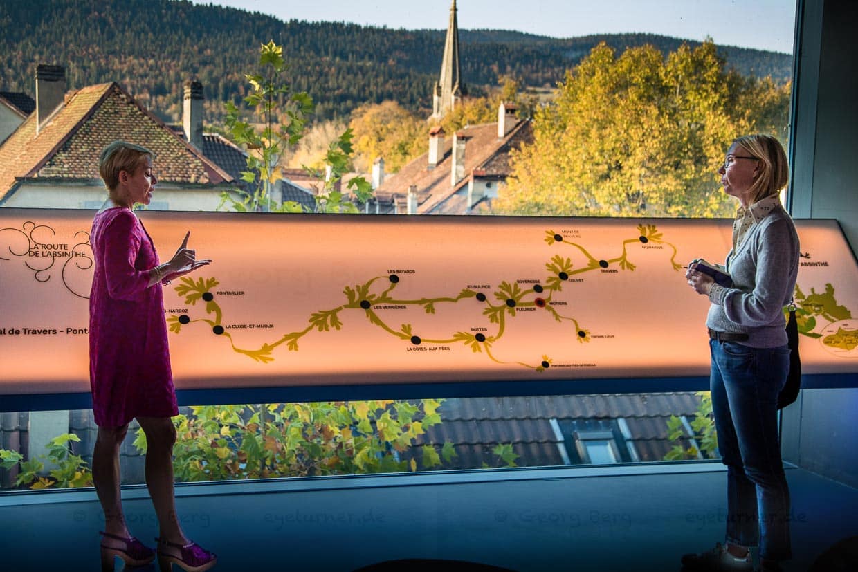 Im Gespräch mit Montse Lucas. Die Spanierin führt sehr kompetent durch die vielseitige Ausstellung im Maison de Absinthe von Motiers. Hier stehen wir vor einer Tafel, die die Route des Absinth zeigt. Eine Wanderstrecke von nur 48 Kilometern verbindet das französische Pontarlier mit dem schweizerischen Noiraigue. Auf der Route liegen zahlreiche Destillerien, Brasserien, Gasthöfe, Museen, Burgen, Hotels und Restaurants, die sich dem Thema Absinth annehmen oder seine Geschichte mitgeprägt haben / © Foto: Georg Berg
