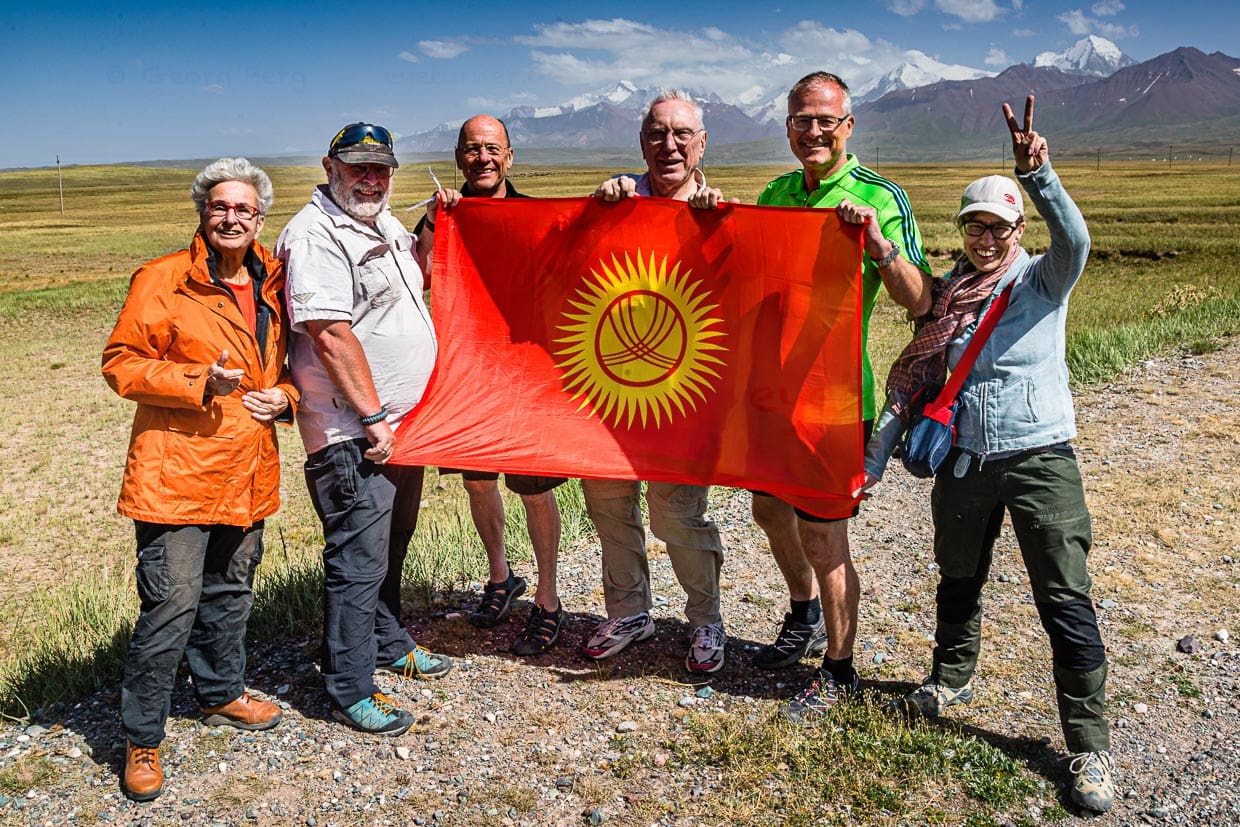 Hinter der kirgisischen Flagge gruppieren sich vor dem Pamir-Gebirge drei Einzelreisende und ein Ehepaar um Veranstalter Konrad Fobbe (2. v.l.) / © Foto: Georg Berg