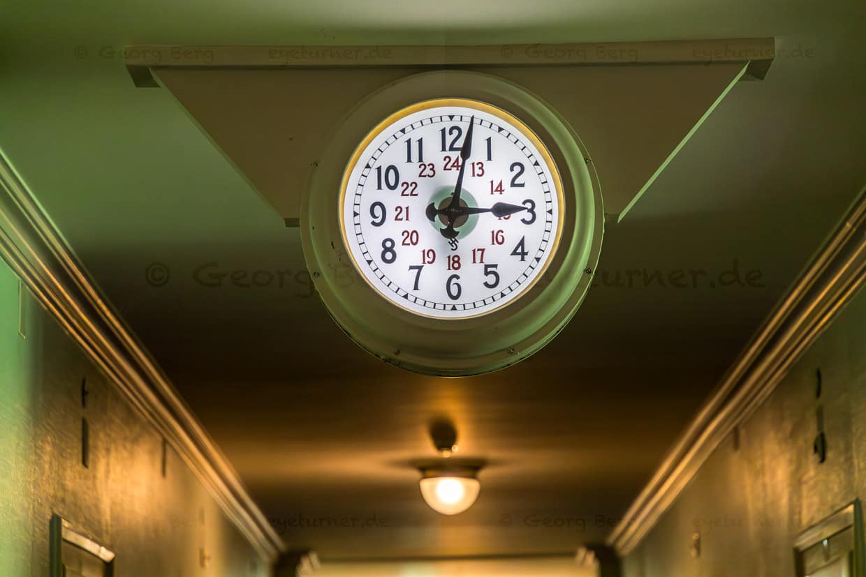 Uhr in den Wohnfluren des ehemaligen Sanatoriums Schatzalp. Modell von Siemens um 1900. Die Uhren, so heisst es, gingen bewusst einige Minuten vor, damit die Patienten ihre Termine, insbesondere die Abfahrt der nahen Schatzalp-Standseilbahn, nicht verpassten. Die auffälligen, gut sichtbaren Uhren sind somit ein Relikt aus der Zeit der Tuberkuloseheilstätte und stehen für die durchgetaktete Organisation des Sanatoriumsalltag / © Foto: Georg Berg