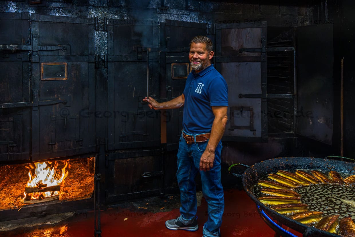Matthias Föh, Firmeninhaber Fischräucherei Föh in vierter Generation, vor den drei Räucheröfen. Geräuchert wird auf Buchen- und Erlenholz, Die Öfen stammen aus den 1920er Jahren und sind mit drei ikonischen Schornsteinen aus Backstein verbunden / © Foto: Georg Berg