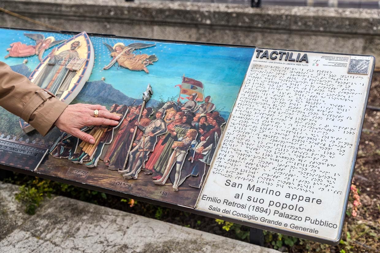 Tactilia in San Marino ist ein taktiles Freilichtmuseum, das 2019 eingeweiht wurde und sich gezielt an Menschen mit Sehbehinderungen oder Blindheit richtet. Es ist Teil des Projekts San Marino Accessible Tourism, das kulturelle Teilhabe für Menschen mit sensorischen oder motorischen Einschränkungen fördert. Gezeigt werden Modelle bekannter Bauwerke, Kunstwerke und historische Symbole. Hier: taktile Tafel über das Gemälde von Emilio Retrosi (Der heilige Marinus erscheint seinem Volk), das im Pallazo Publico hängt / © Foto: Georg Berg