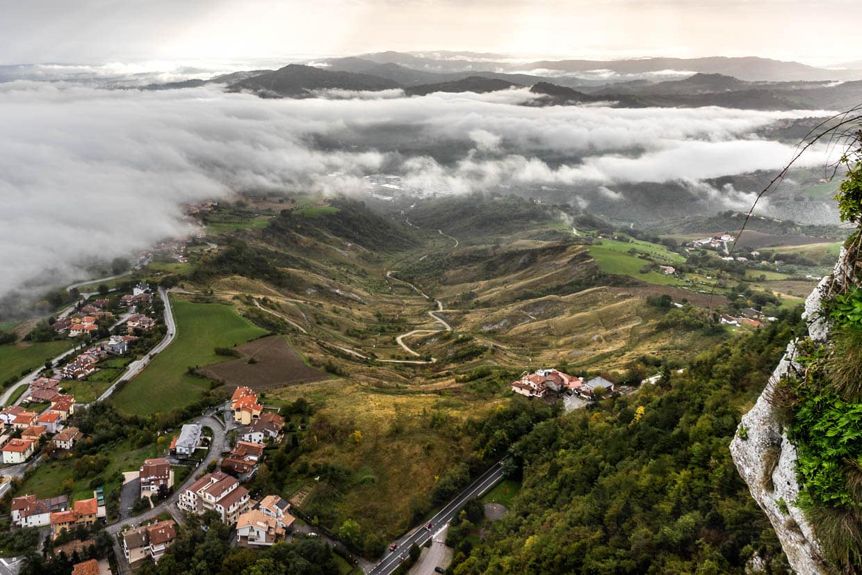 Blick vom Monte Titano auf einen Industriekomplex am Ende des Tales. Dort hinter den Wolken befindet sich auf der abgewandten Seite das Wandgemälde "Die Geschiche von San Marino" des brasilianischen Künstlers Eduardo Kobra / © Foto: Georg Berg