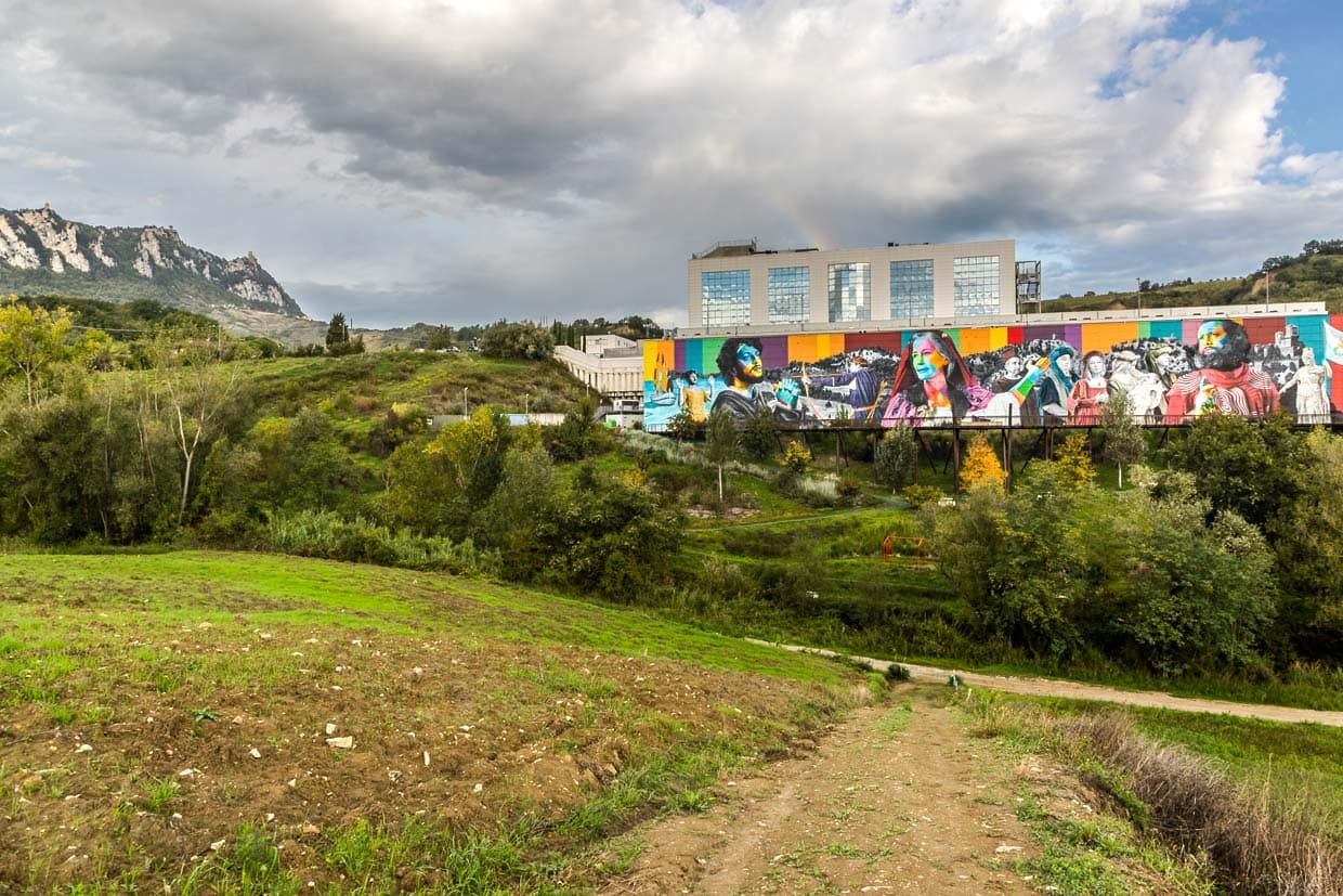 Ein Wandgemälde des brasilianischen Künstlers Eduardo Kobra trägt den Titel "Die Geschichte San Marinos". Die farbenprächtige Darstellung historischer Persönlichkeiten und Szenen ist mitten in der hügeligen Landschaft San Marinos auf der Fassade einer Verpackungsfabrik angebracht. Im Hintergrund auf dem Monte Titano die drei ikonischen Türme im historischen Zentrum von San Marino. / © Foto: Georg Berg
