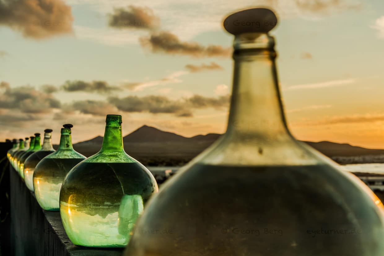 Reihe von grünen Weinballons auf Lanzarote bei Sonnenuntergang. Bei der traditionellen teiloffenen Gärung schützt ein Stein den Wein vor Verunreinigungen / © Foto: Georg Berg