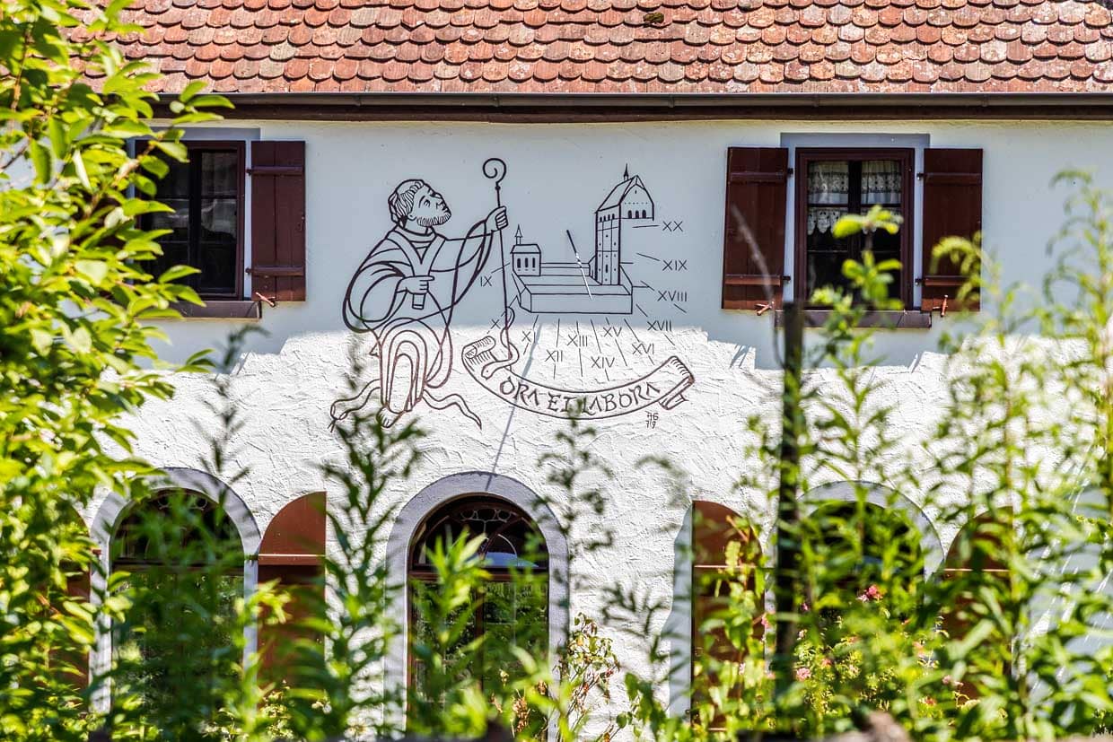 Hausfassade nahe dem Münster St. Maria und Markus in Mittelzell mit der Darstellung eines Benediktinermönches und dem Ordensspruch Ora et Labora. Auf der Insel Reichenau galt die Ordensregel „Ora et labora“ seit der Gründung 724 die Mönche: Sie beteten im Münster und bewirtschafteten Gemüsefelder sowie Weinberge, was die Insel zur „Gemüse- und Weininsel“ machte. Das Motto unterstreicht die UNESCO-Welterbe als Symbol für harmonisches Mönchsleben / © Foto: Georg Berg