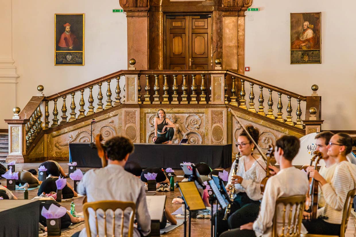 Eine Frau posiert auf einer Bühne während Musiker spielen und Menschen Yoga praktizieren am Domplatz Salzburg Austria