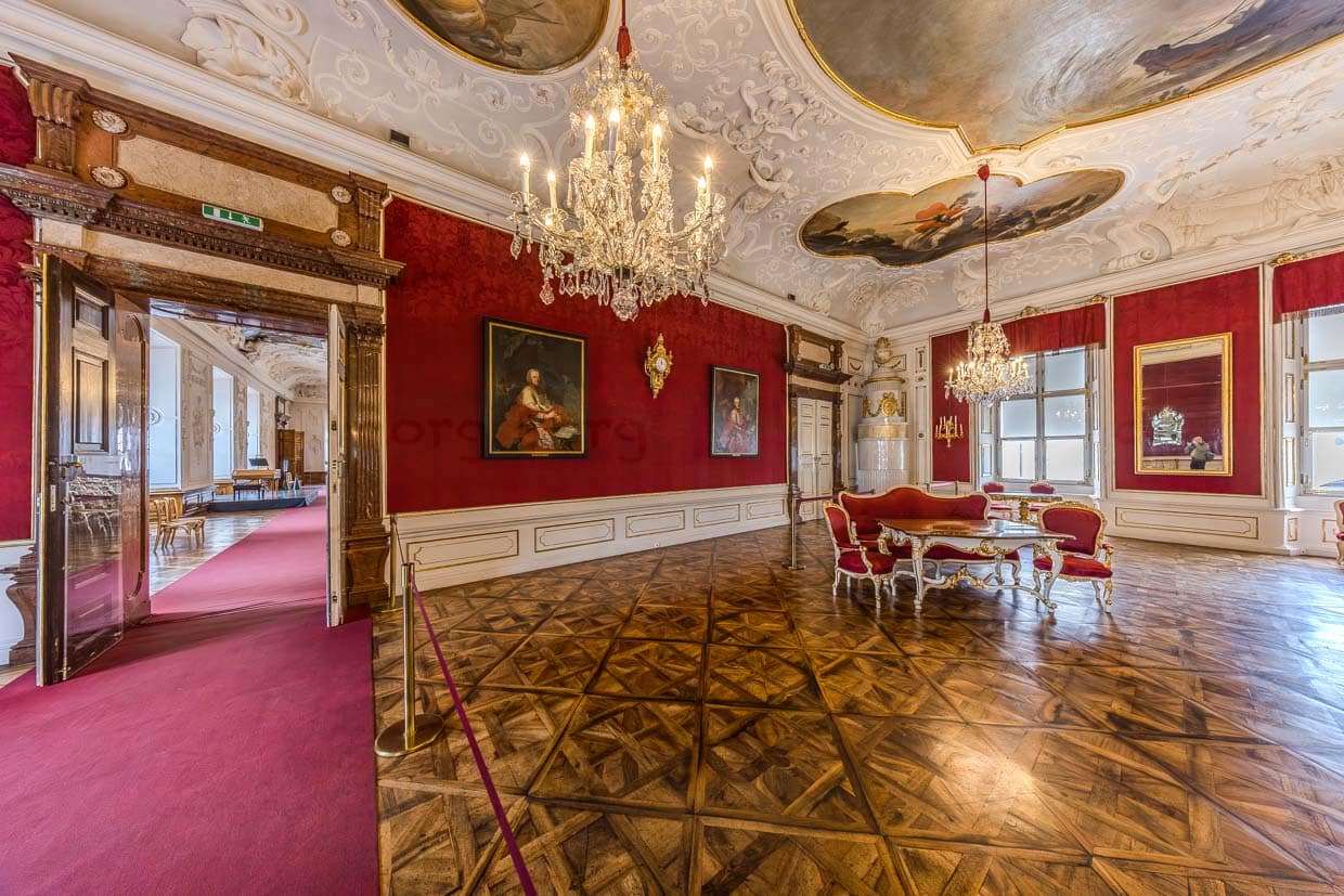 Ein opulenter Barocksaal mit roten Wänden, Deckenmalereien, Kronleuchtern und antiken Möbeln zeigt sich am Residenzplatz in Salzburg Austria