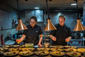 Mit Standfestigkeit und ruhiger Hand: Martina und Gavin richten in der Kombüse trotz Seegang das Hauptgericht an / © Foto: Georg Berg