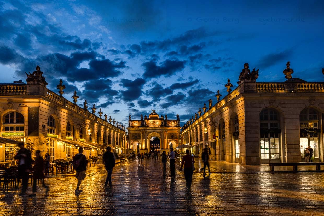 Menschen spazieren über den nassen Platz Stanislas in Nancy, Frankreich, bei Einbruch der Dunkelheit