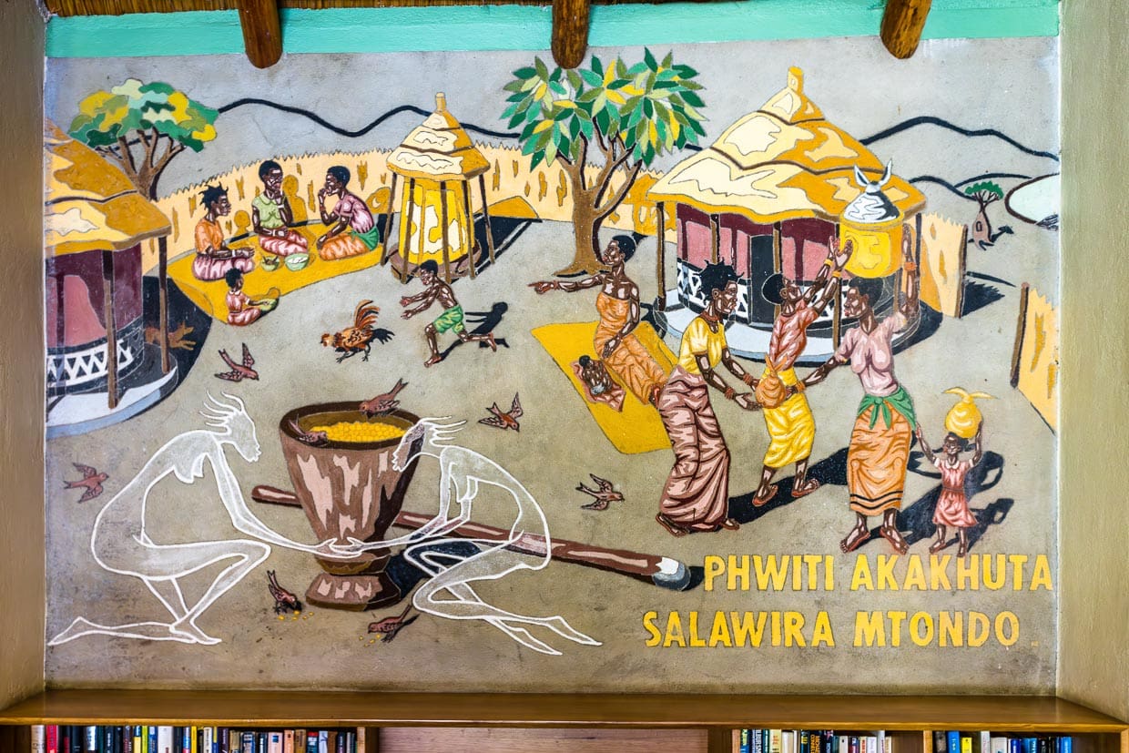 Fresko Malawian Hospitality im Makokola Retreat am Lake Malawi. Das Bild zeigt wichtige Bestandteile der Gastfreundschaft. Darunter das Mahlen von Mais für das Nationalgericht Nsima. Vorne links großer Mörser mit Stößel / © Foto: Georg Berg