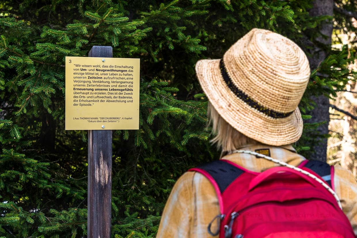 Spaziergängerin liest Texttafel mit Zitaten aus „Der Zauberberg“ auf dem Thomas-Mann-Weg in Davos. Der Weg führt vom ehemaligen Waldsanatorium (heute Waldhotel Davos) auf etwa 1.620 m Höhe bis zur Schatzalp auf 1.880 m Höhe / © Foto: Georg Berg