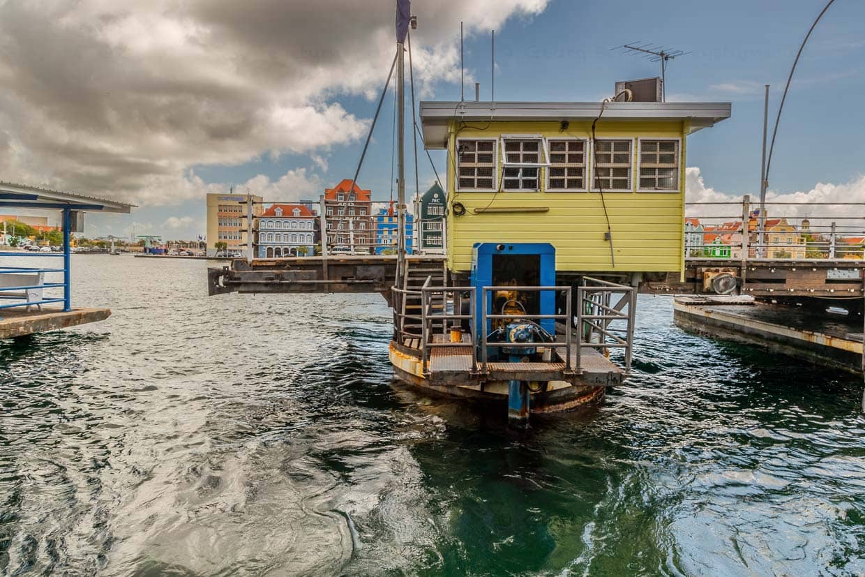Im geöffneten Zustand ist der äußere Ponton mit dem Führerhaus, der im geschlossenen Zustand der Brücke am Punda-Ufer liegt, vollständig bis zum Ufer von Otrabanda gefahren. Links im Hintergrund ist die Lloyd Benjamin Smith Brücke zu erkennen, die das Schottegat zum kleinen Hafen Waaigat überquert / © Foto: Georg Berg