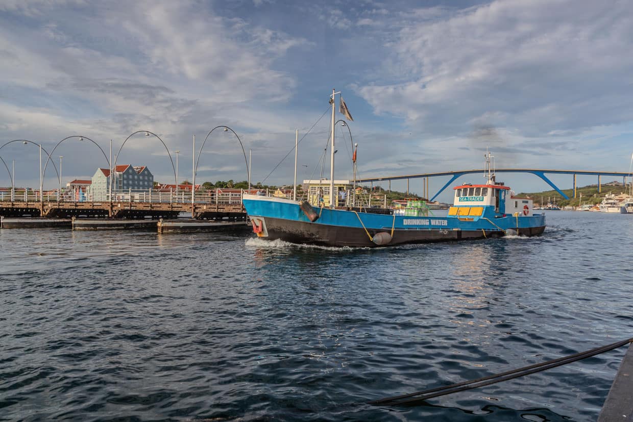 Ein Trinkwasserschiff aus Curaçao passiert die halb geöffnete Ponton-Brücke von Willemstad. Die Königin-Emma-Brücke wird „Swinging Old Lady“ genannt. Diese Ponton-Fußgängerbrücke aus dem Jahr 1888 öffnet sich seitlich für vorbeifahrende Schiffe. Autos können die St. Anna Bucht seit 1974 über die Königin Juliana Brücke (im Hintergrund) überqueren / © Foto: Georg Berg