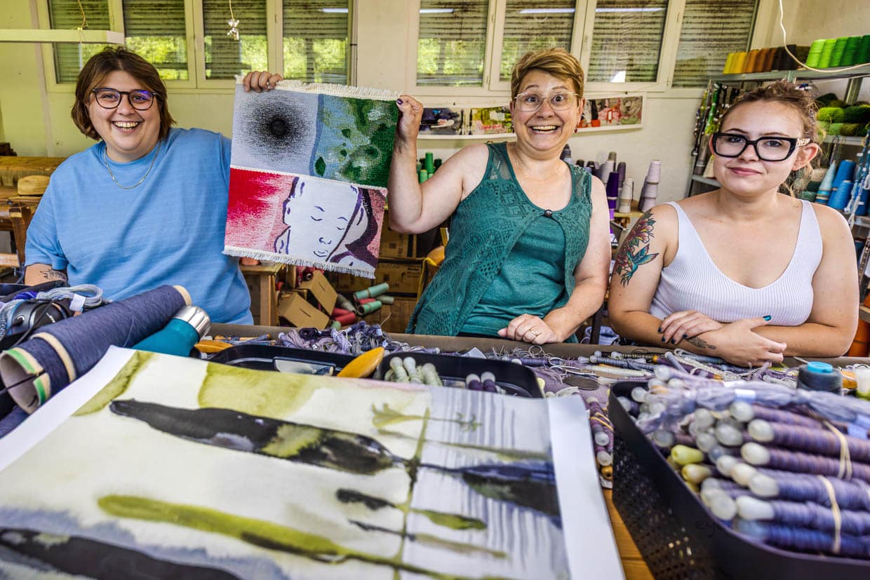 Sarah Chassain, Teamleitung für das Projekt Wandteppich "Hommage à George Sand“ im Atelier Robert Four mit den Kolleginnen Frederique und Agathe. Sie halten das Musterstück hoch, mit dem sie sich für Ausschreibung beworben haben. Das Musterstück zeigt auf kleiner Fläche verschiedenen Fertigkeiten, die ein Team für die Realisierung des Teppichs beherrschen muss / © Foto: Georg Berg